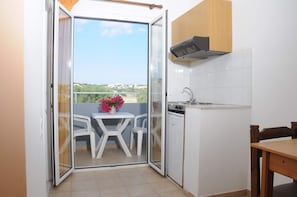 A compact kitchen with a sliding glass door leading to a balcony, a small table with chairs, and a view of the surrounding area. - Mastorakis Studios (Hersonissos)