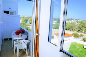 Un balcón con una mesa y sillas blancas, un florero con flores y vistas a un paisaje verde y edificios.