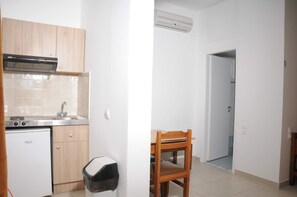 Une cuisine compacte équipée d’une cuisinière intégrée, d’un évier et de placards en bois. On y trouve un petit réfrigérateur et une poubelle. Une chaise en bois est placée près d’une porte-fenêtre donnant sur une autre pièce.