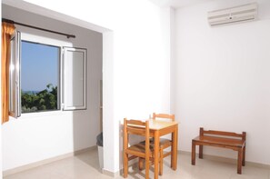 A room with a window, a table with chairs, a bench, and an air conditioning unit. - Mastorakis Studios (Hersonissos)