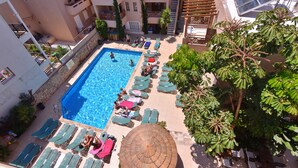 Une piscine sur le toit, dotée de chaises longues et de parasols, entourée d’immeubles et d’arbres.