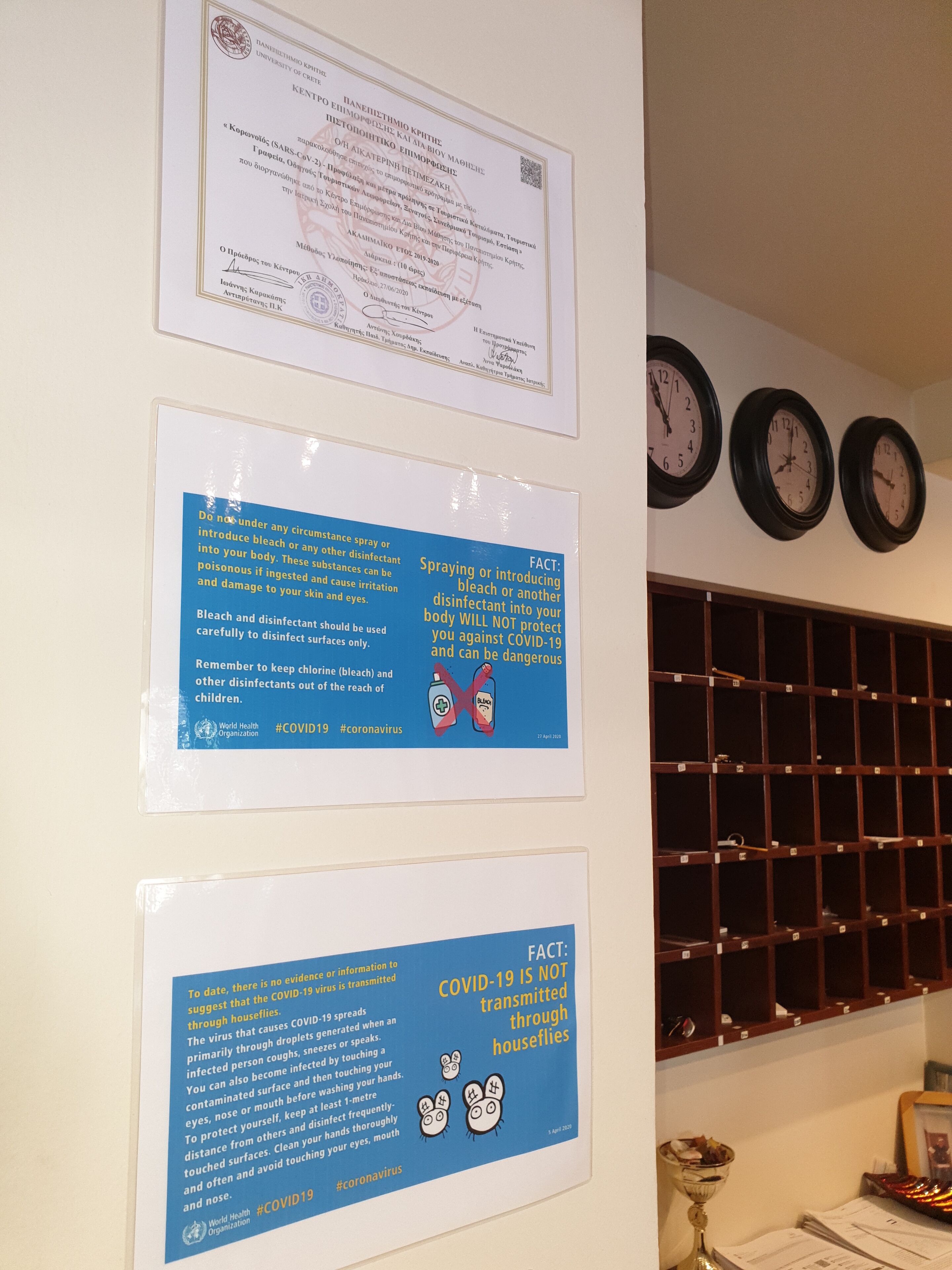 A wall with educational posters about COVID-19, a framed diploma, and three wall clocks.