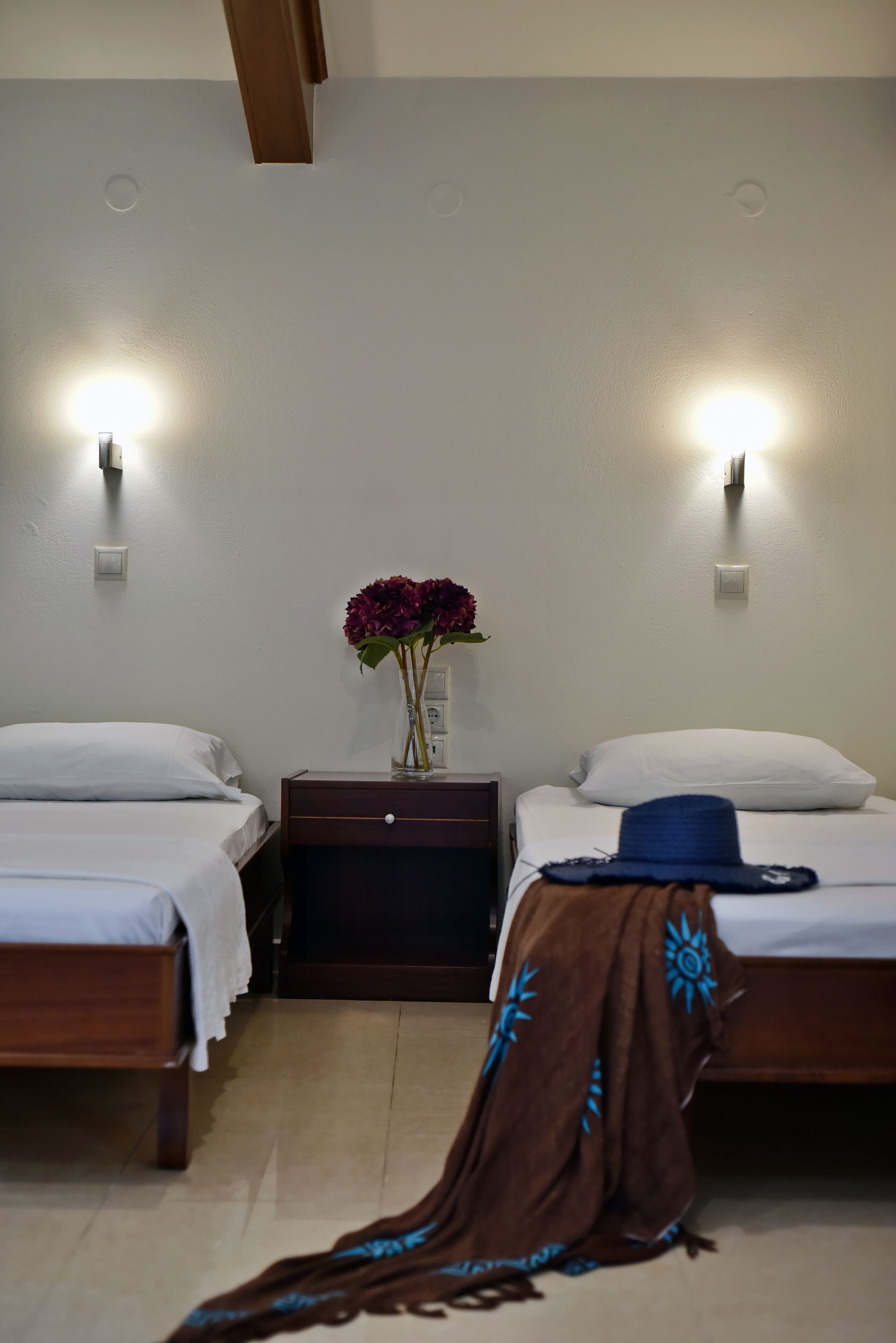 Une chambre d’hôtel avec deux lits, une table de chevet, un vase de fleurs et un luminaire fixé au mur.