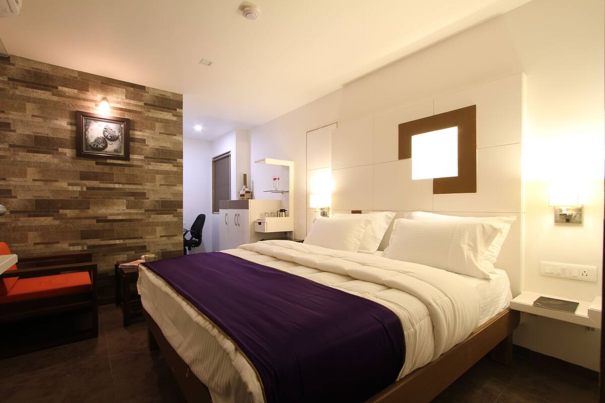 A hotel room with a bed, a desk, a chair, a small table, a wall-mounted shelf, and a picture frame.