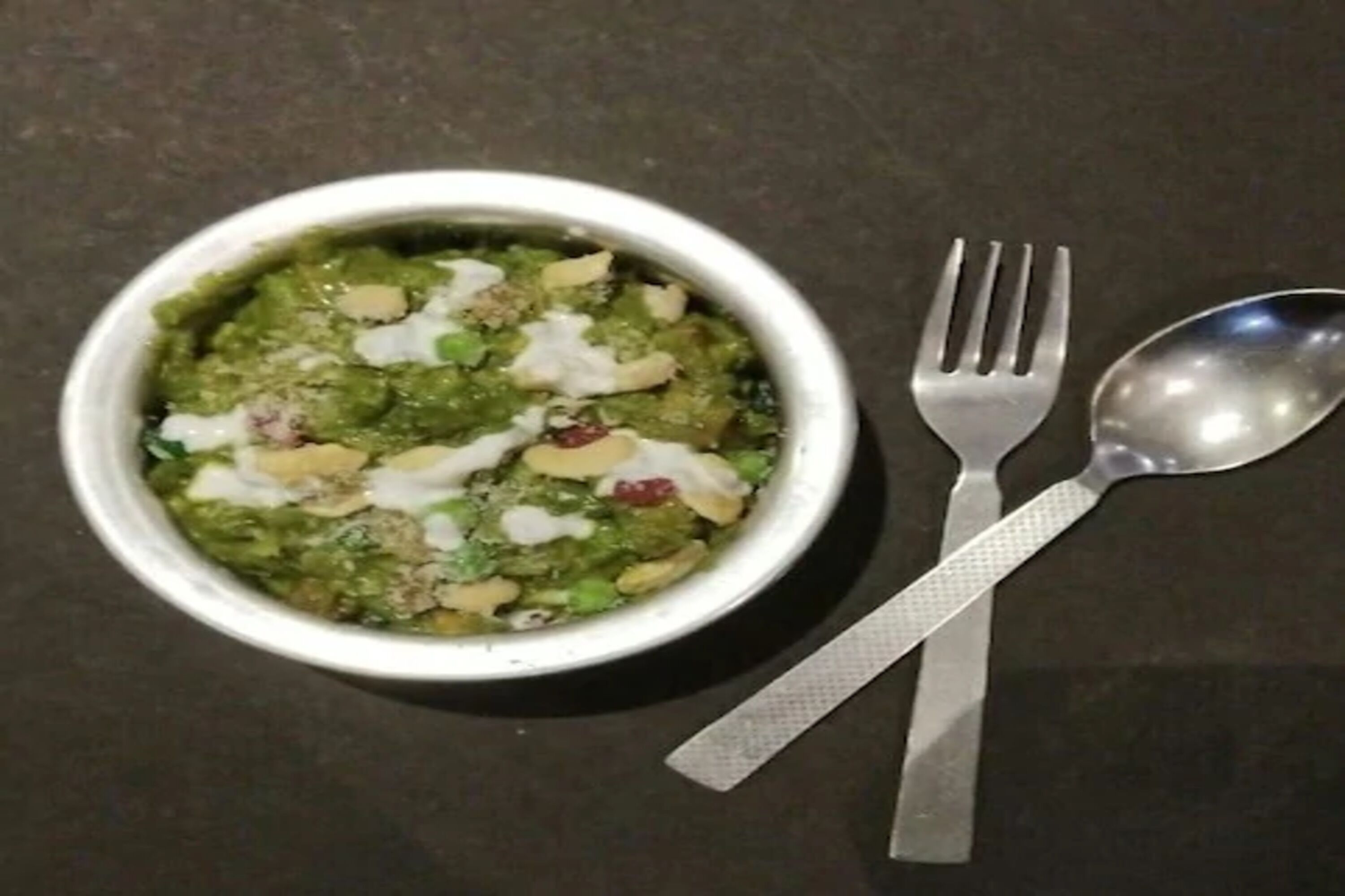 A bowl of green soup with chunks of meat and cheese on a dark surface.