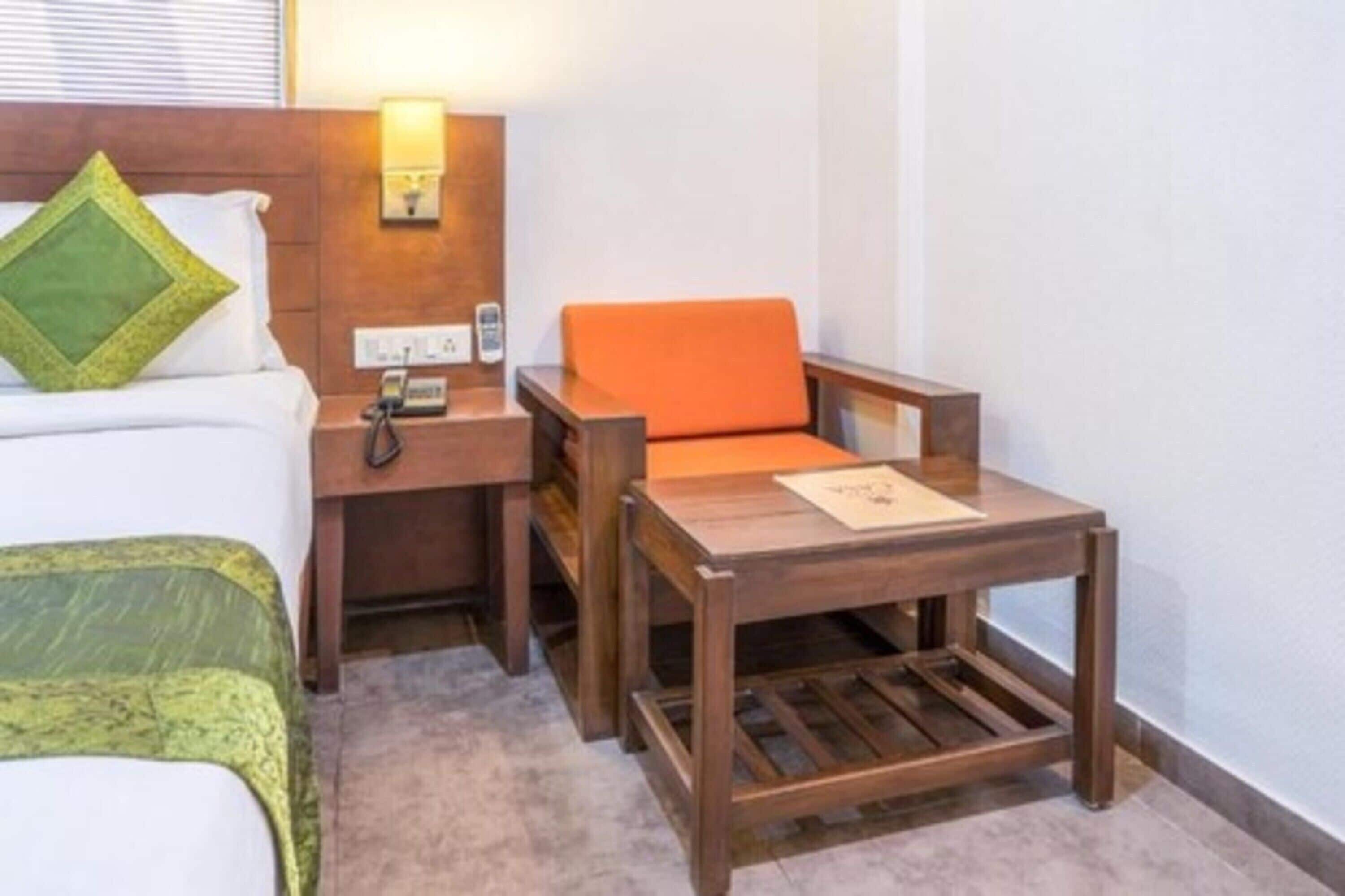 A hotel room with a bed, a nightstand with a lamp, a wooden table, and an orange chair.