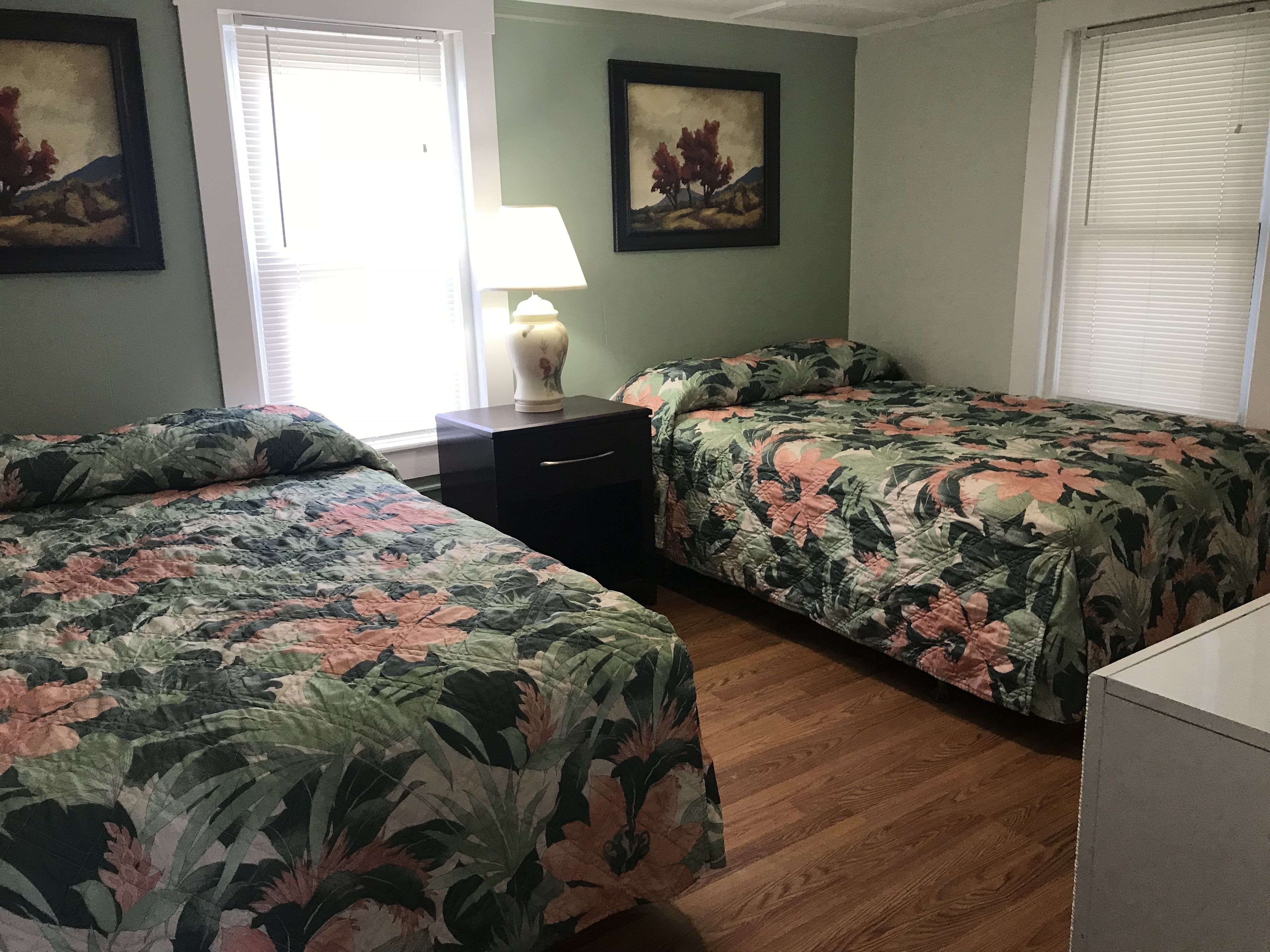 A room with two beds, a nightstand, a lamp, and two framed paintings on the walls.