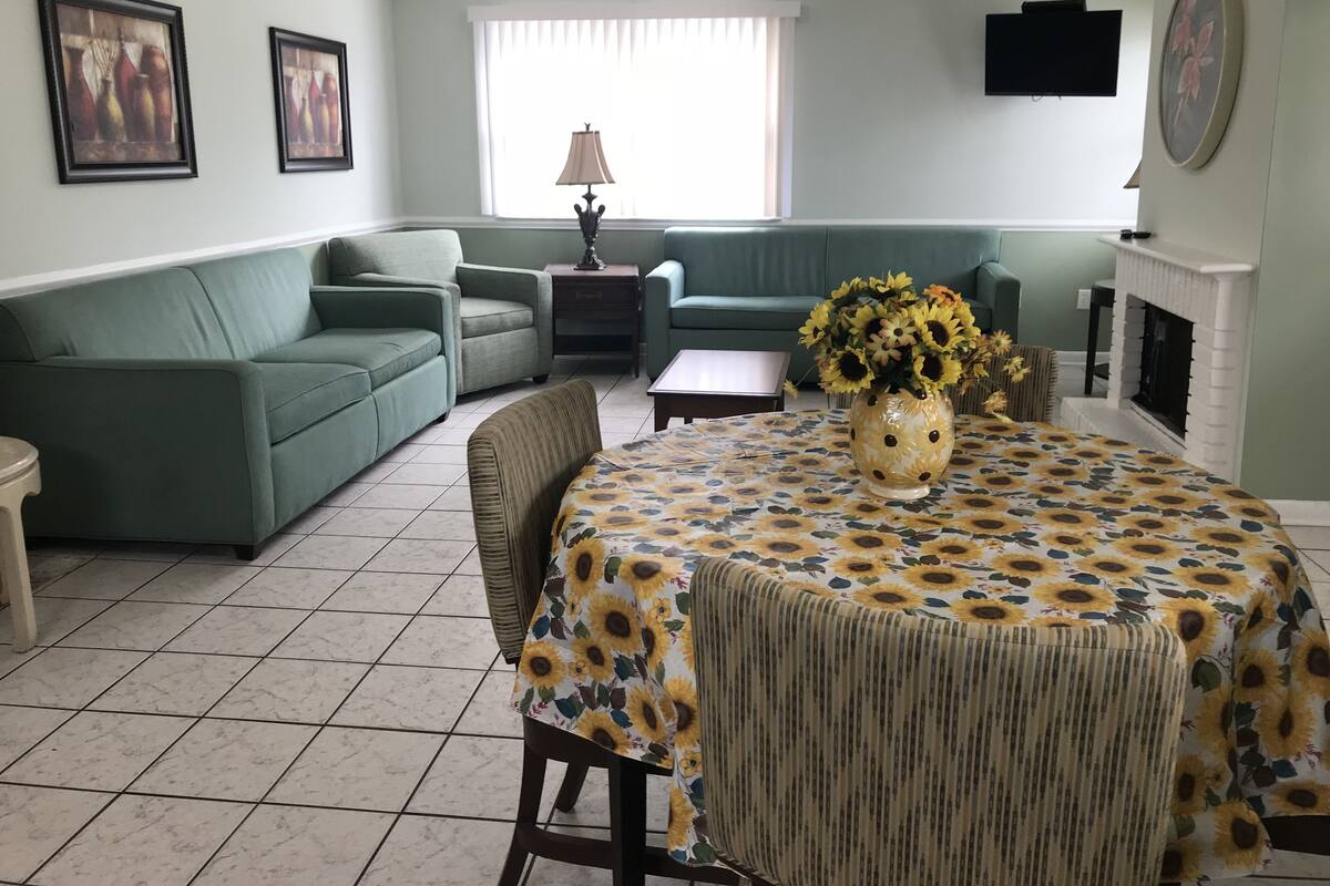 Una sala de estar con un sofá verde, una mesa de comedor con mantel de girasoles y una chimenea.