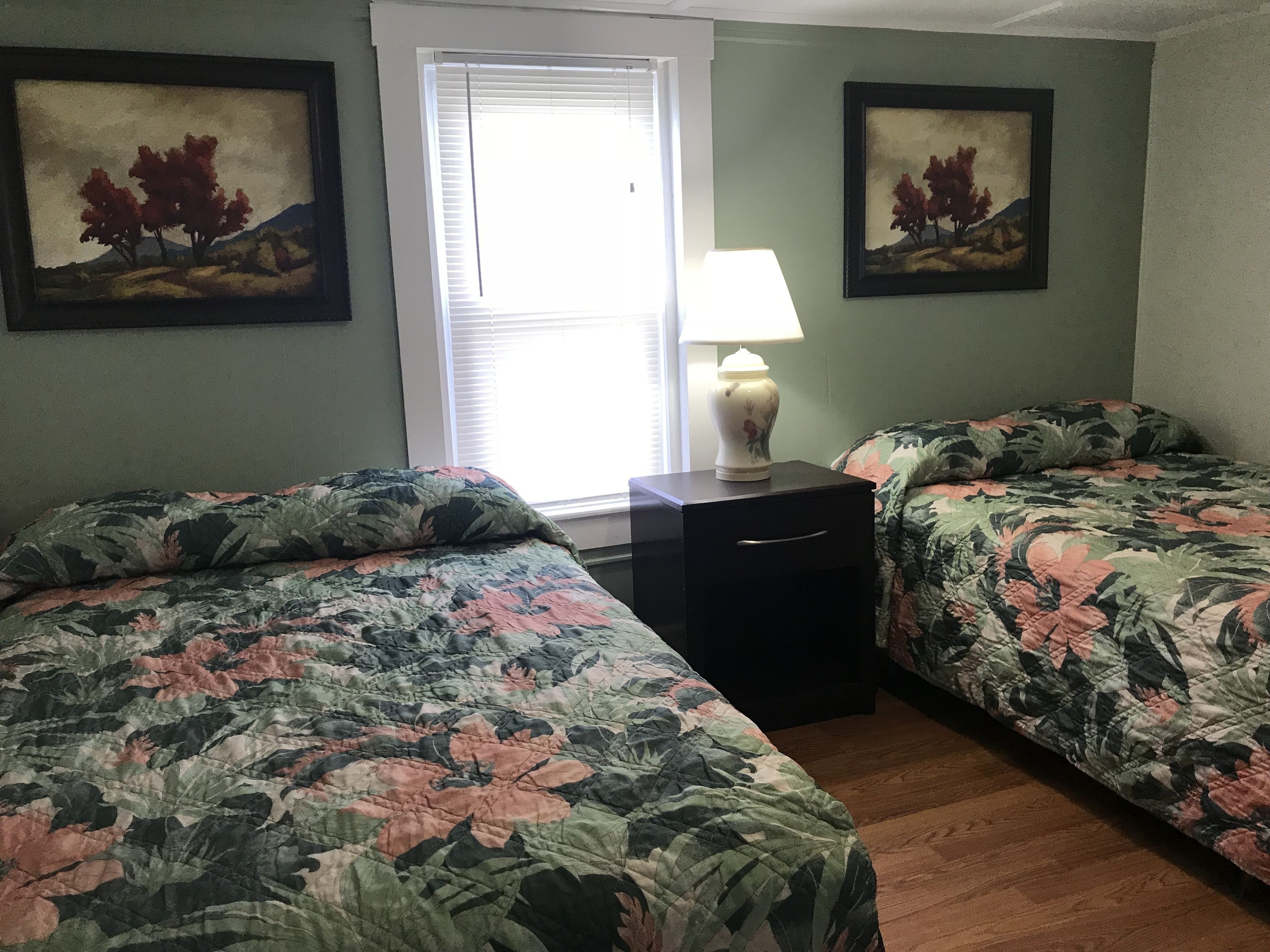 A room with two beds, a nightstand, a lamp, and two paintings on the walls.