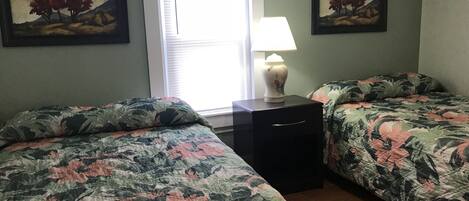 A room with two beds, a nightstand, a lamp, and two paintings on the walls.