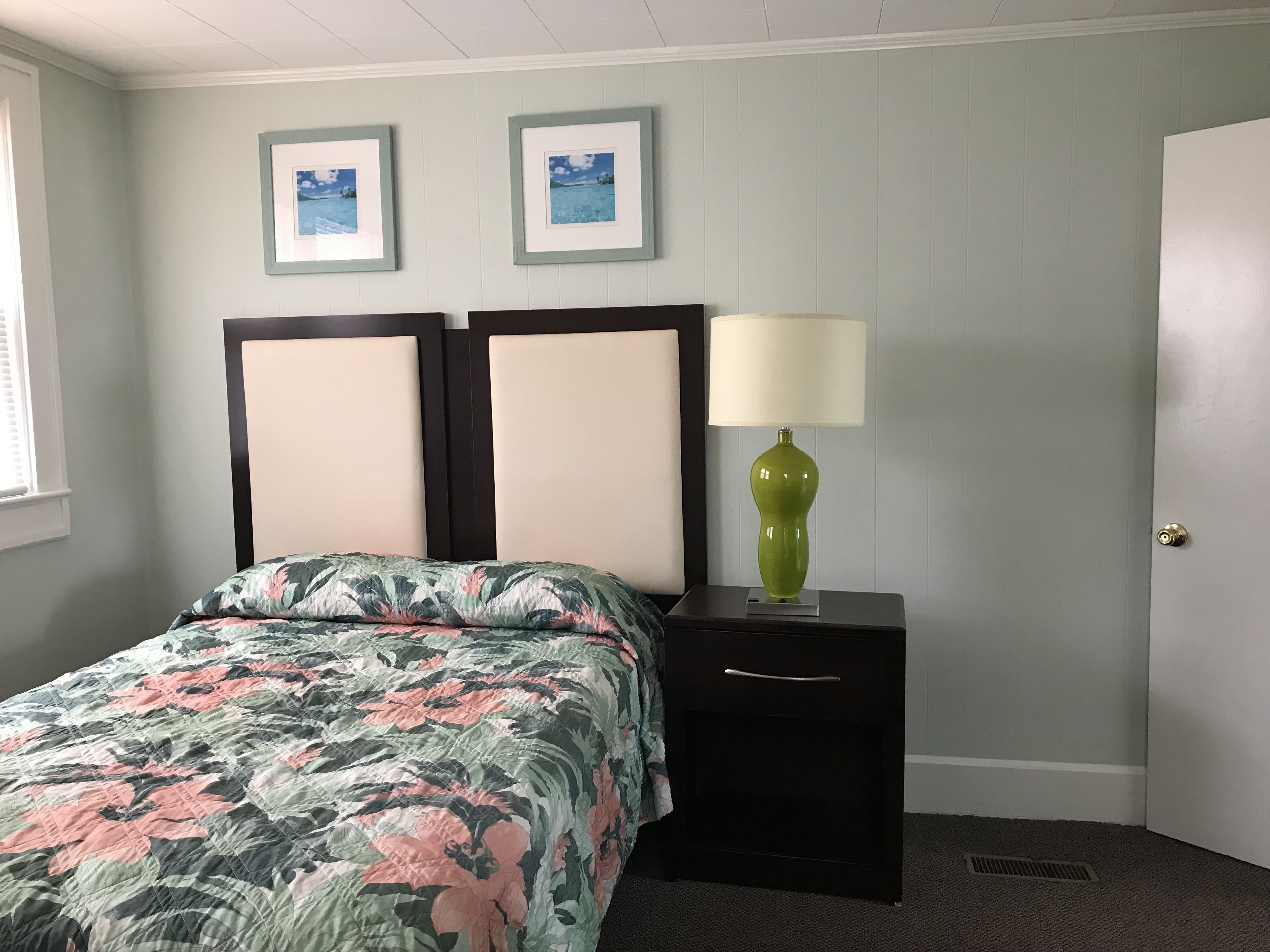 A bedroom with a floral bedspread, a nightstand with a green lamp, and two framed pictures on the wall.