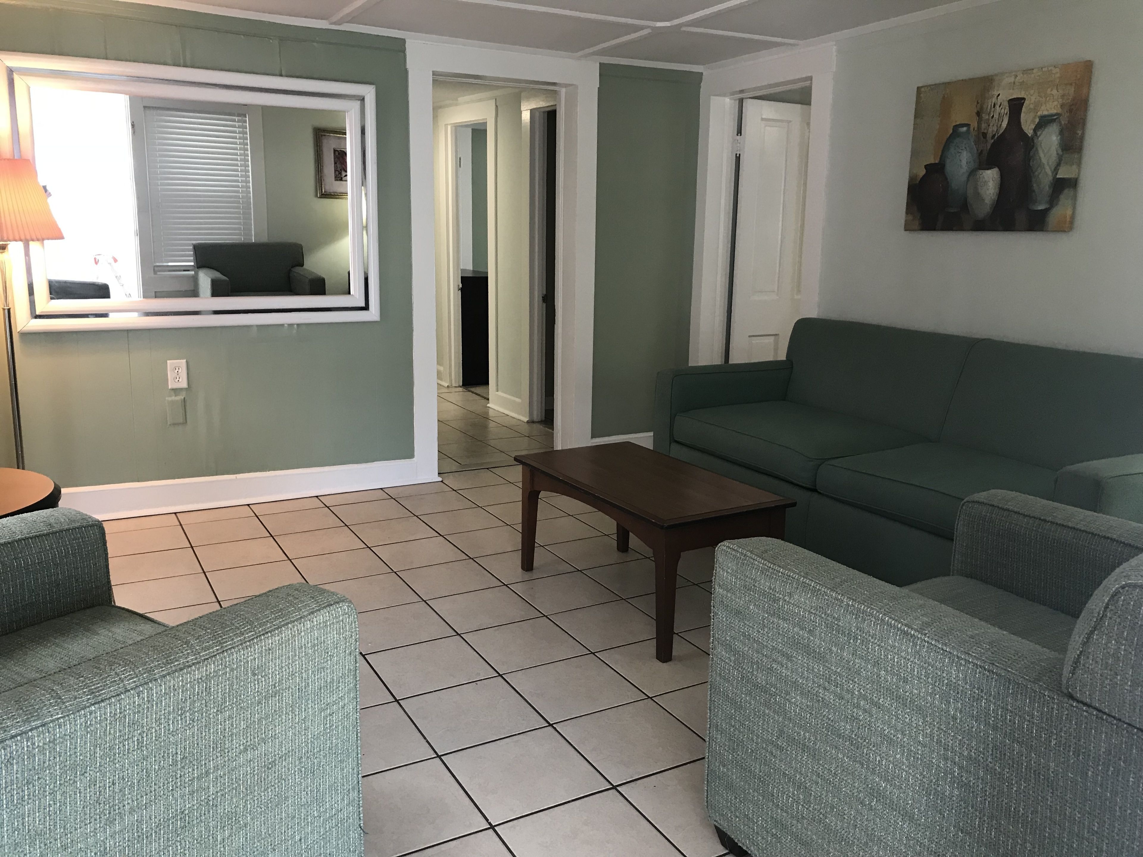 Ein Wohnzimmer mit einem grünen Sofa, einem kleinen Holztisch und zwei Sesseln. An der Wand hängt ein Gemälde und es gibt ein Fenster mit Jalousien.