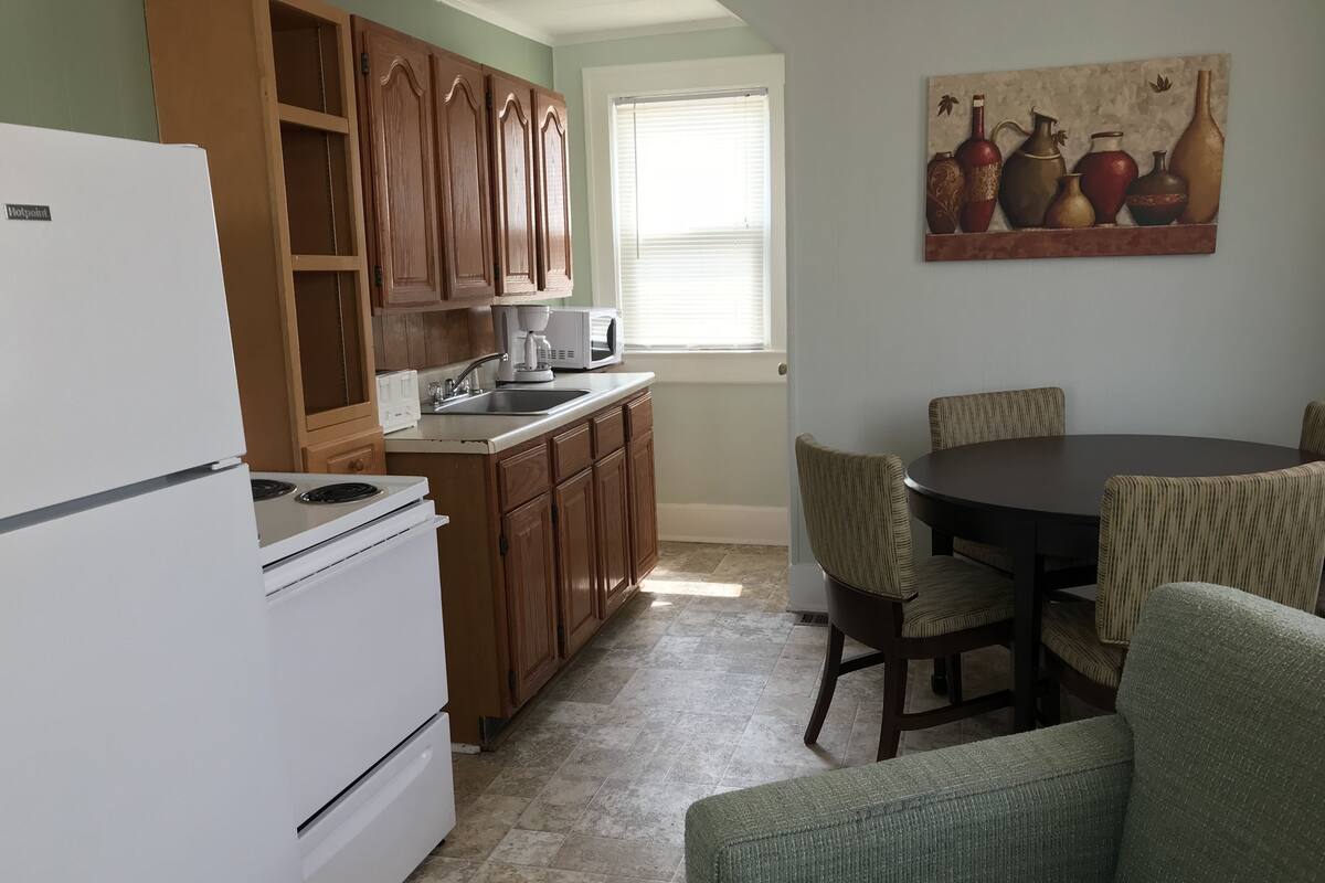 Una cocina compacta con armarios de madera, un refrigerador blanco, un microondas y un comedor con mesa y sillas.