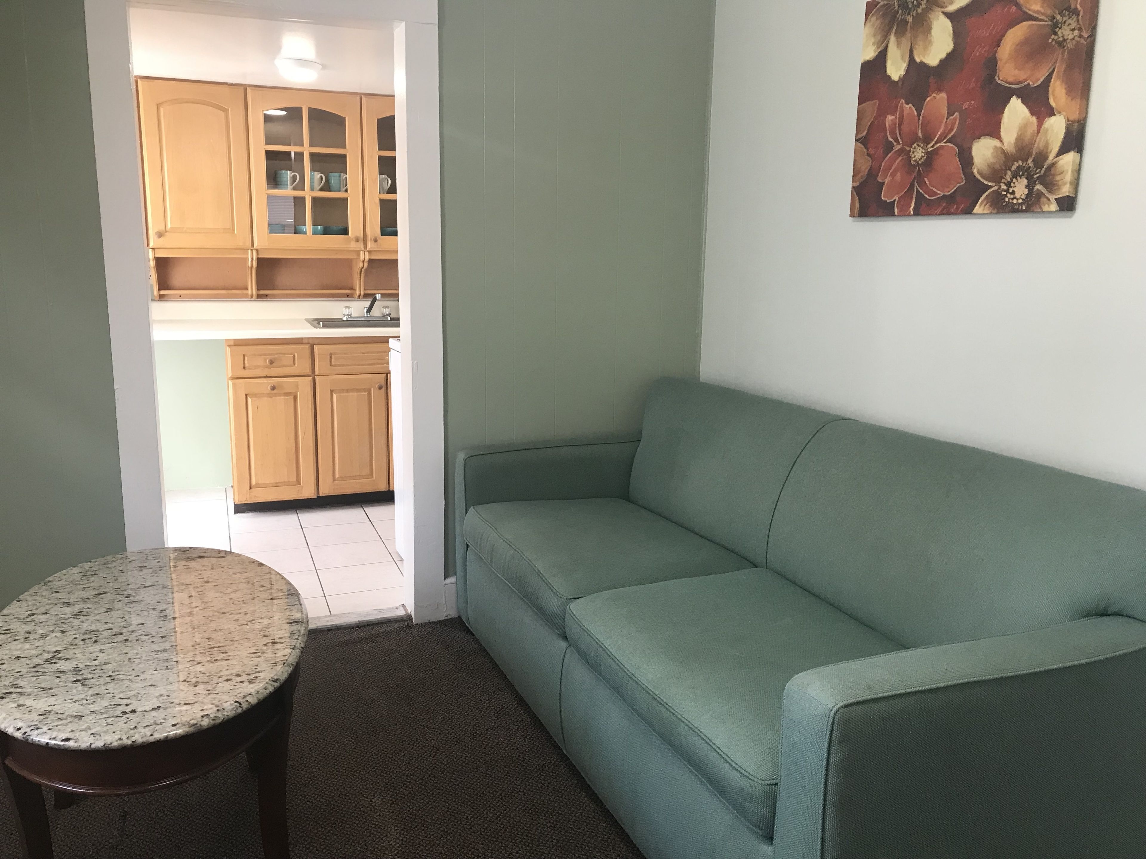 A living room with a green sofa, a round table, and a kitchen in the background.
