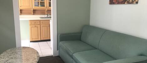 A living room with a green sofa, a round table, and a kitchen in the background.