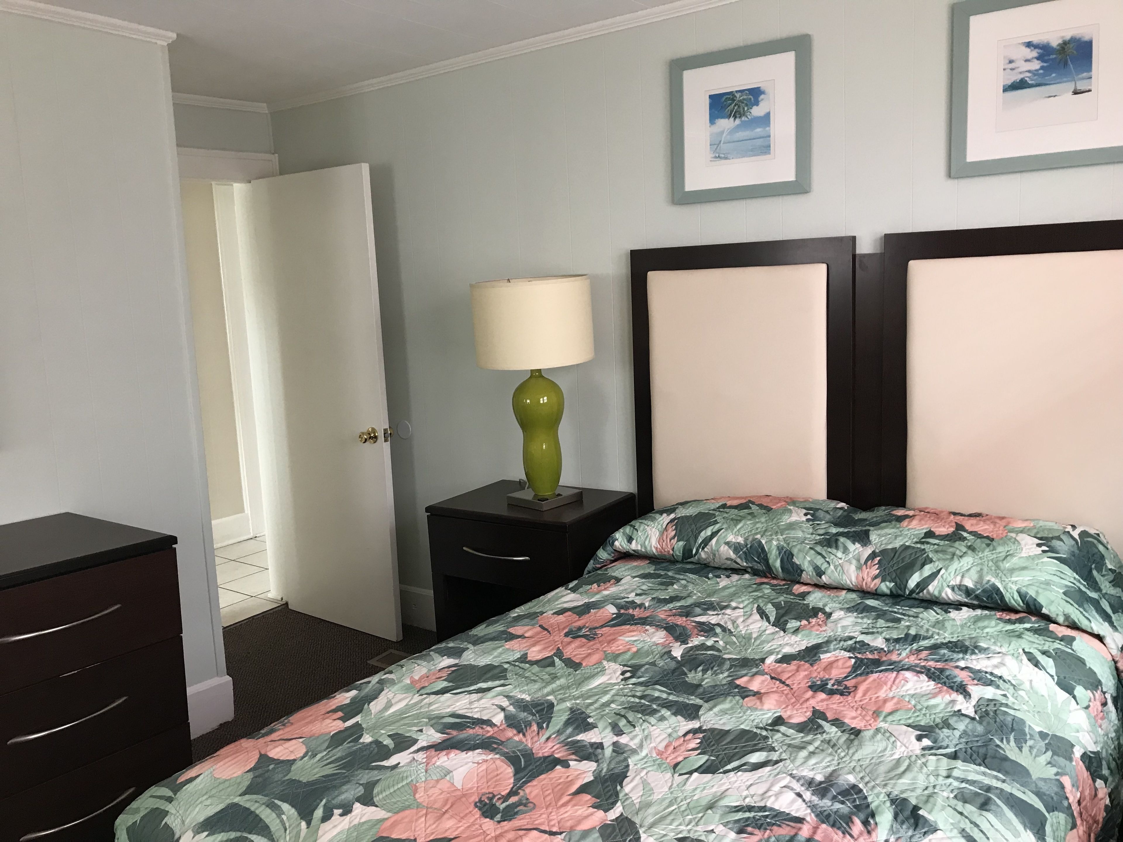 Hotellihuone, jossa on sänky, yöpöytä lampun kanssa, lipasto ja kaksi kehystettyä kuvaa seinällä.