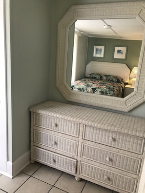 Un dormitorio en esquina con un espejo grande, una cama con ropa de cama floral, un aparador de mimbre y una mesita de noche con lámpara.