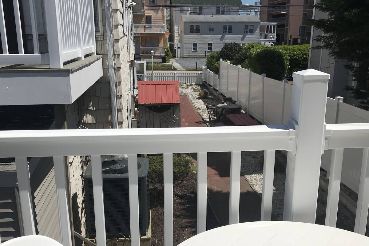Un balcón con sillas y una mesa blancos, con vista a una calle con edificios y una zona cercada.