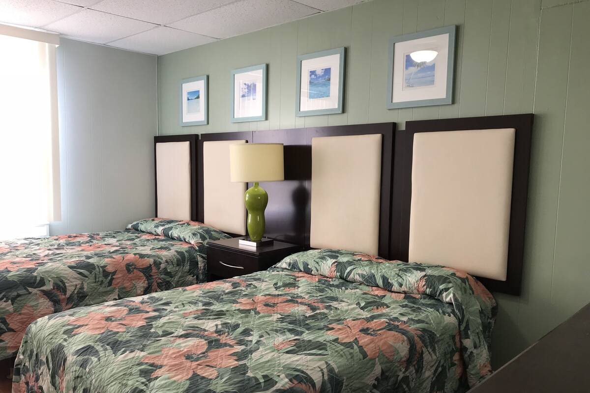 Habitación de hotel con dos camas, una mesita de noche, una lámpara y cuadros en la pared.