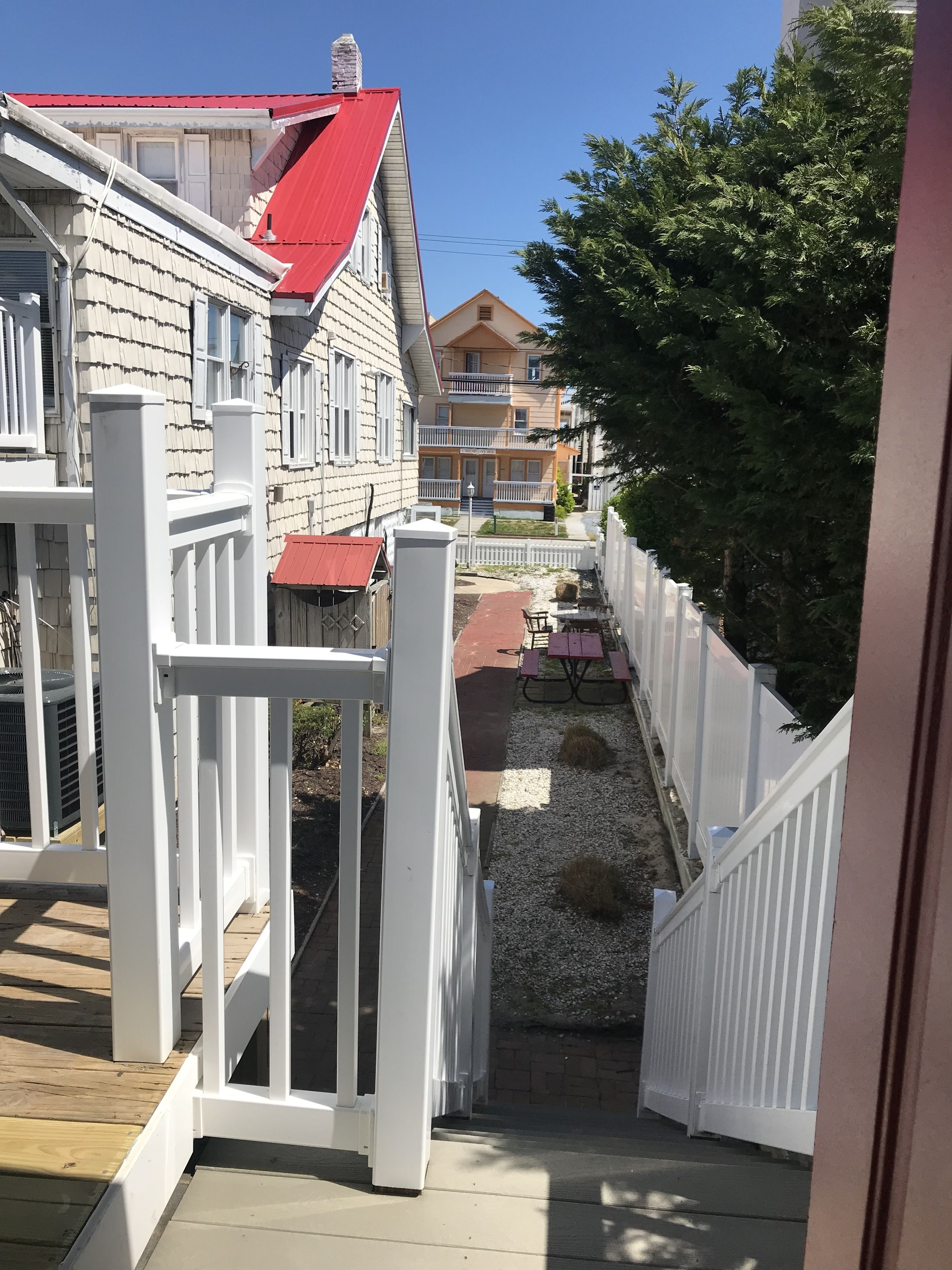 A wooden deck with a white railing, a red roof, and a gravel path leading to a picnic table and benches.