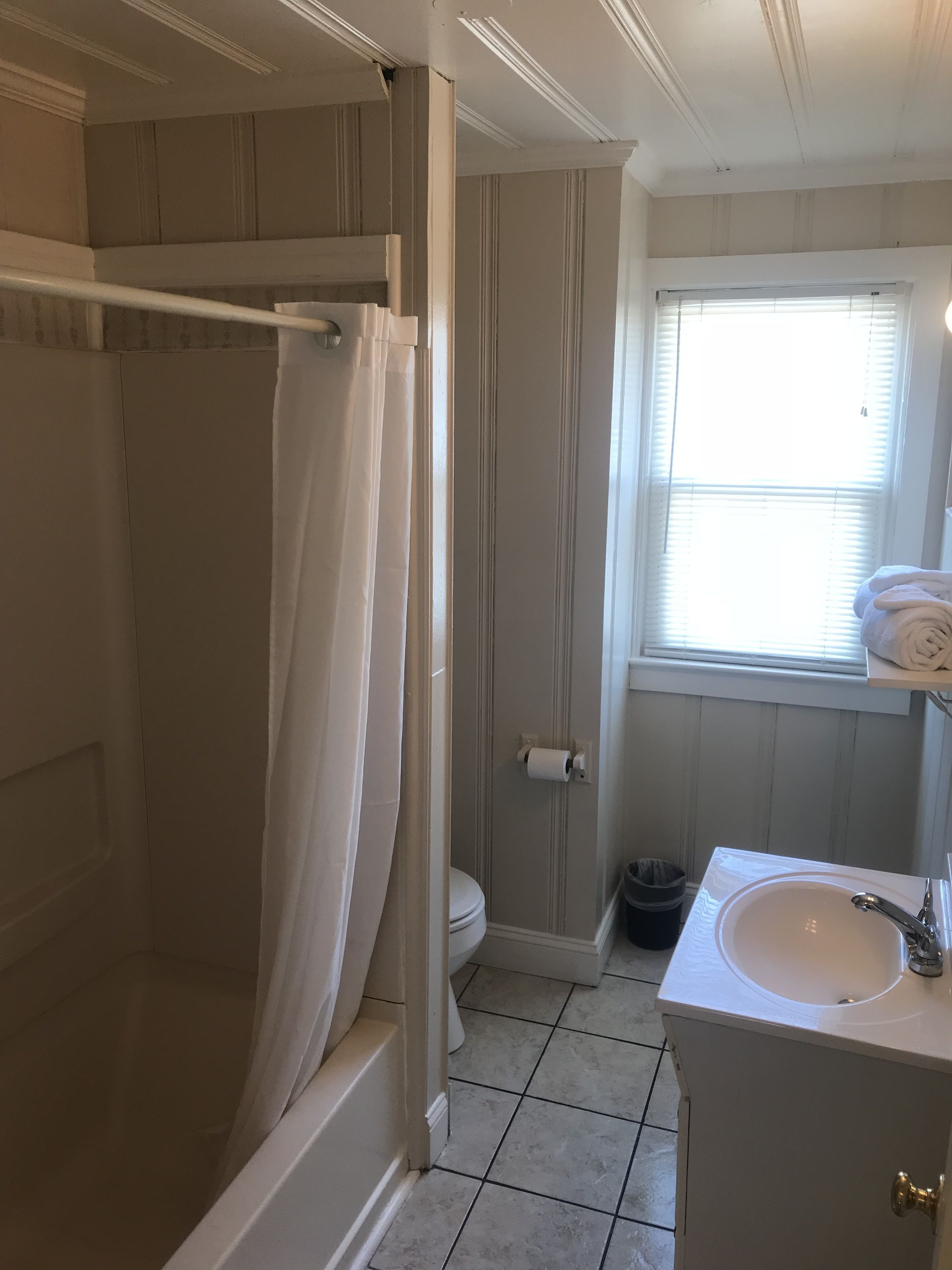 Un baño con ducha, inodoro, lavamanos y una ventana con persianas.