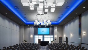 Un salón de conferencias con filas de sillas orientadas hacia una pantalla grande, un atril y un techo iluminado en azul.