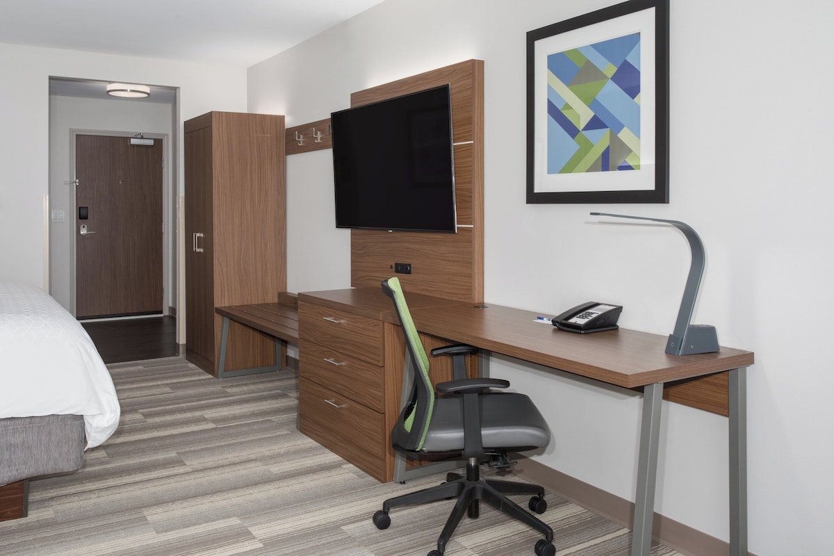 A hotel room with a bed, a desk with a chair, a television, a framed picture, and a desk lamp.