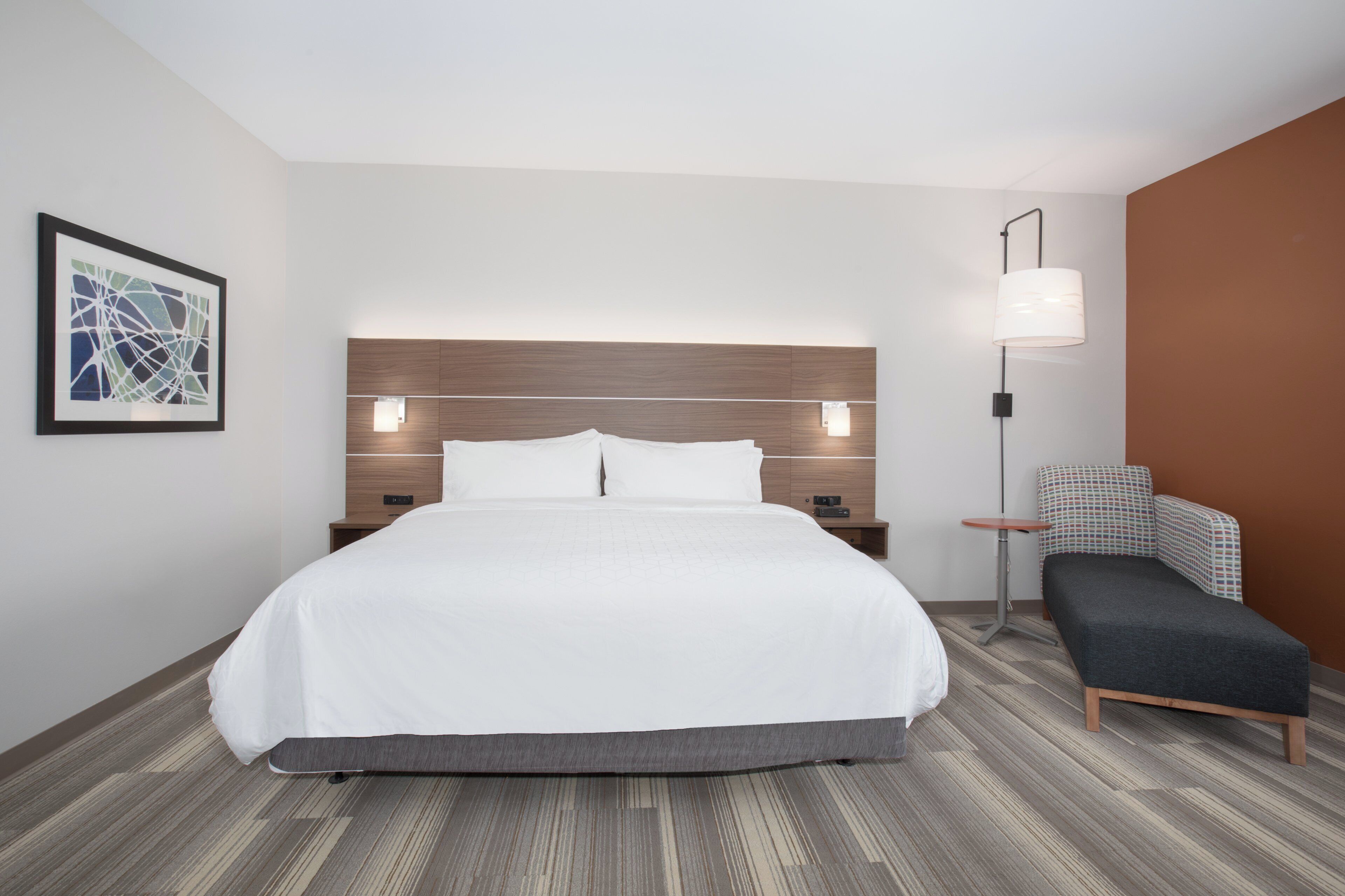 A hotel room with a large bed, a bedside table, a chair, a lamp, and a framed picture on the wall.