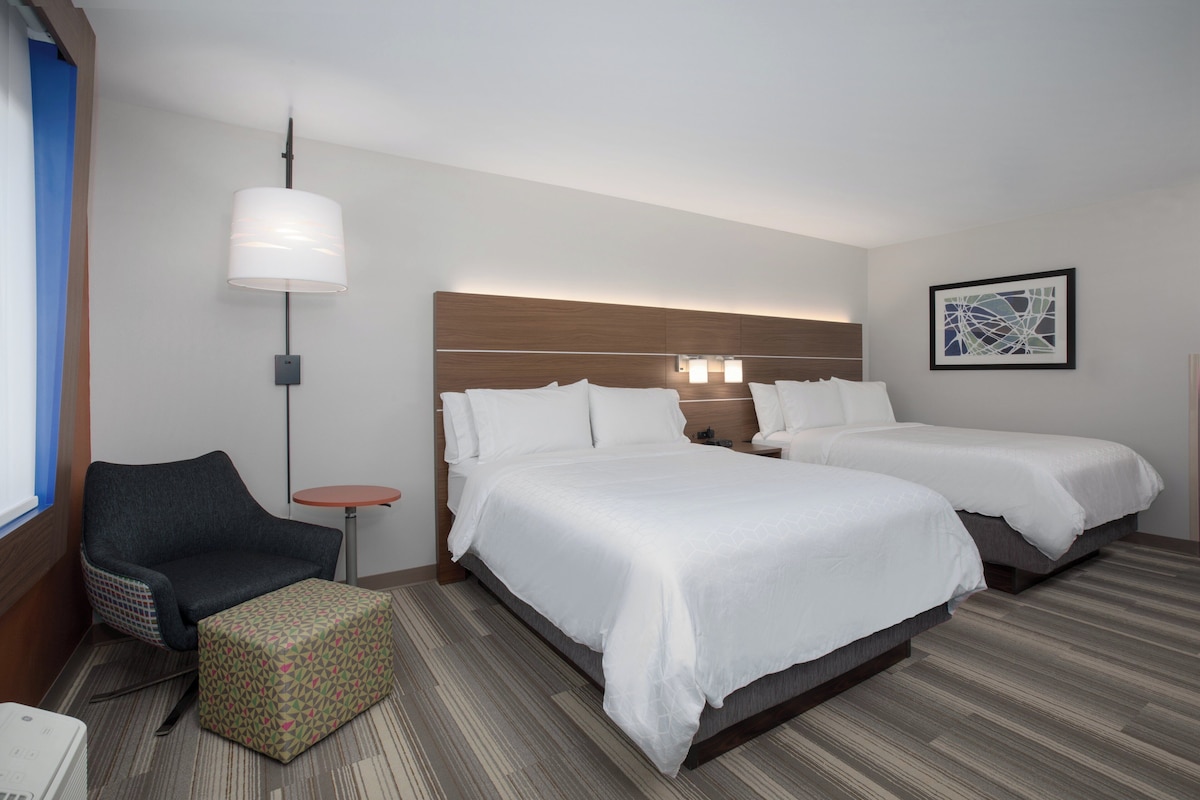 A hotel room with two beds, a chair, a small table, and a wall-mounted lamp.