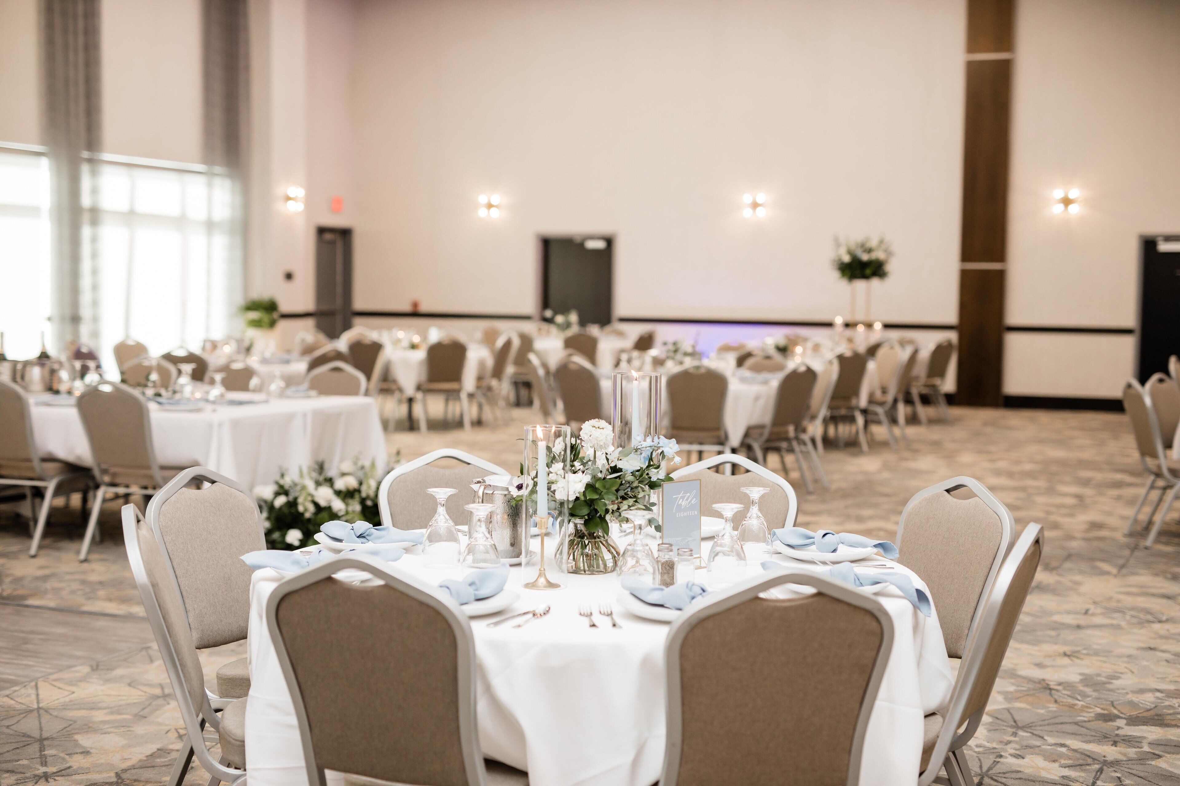 Une salle de banquet avec des tables rondes dressées pour un événement formel, agrémentée de nappes blanches, de compositions florales et de vaisselle élégante.