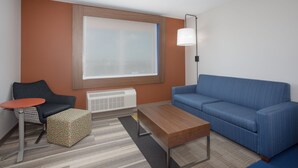Una habitación de hotel moderna con un sofá azul, una mesa de centro de madera, un taburete estampado, una pequeña mesa redonda y un gran ventanal con persianas.