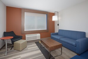 A modern hotel room with a blue sofa, a wooden coffee table, a patterned ottoman, a small round table, and a large window with blinds. - Holiday Inn Express & Suites Sioux City North-Event Center by IHG (North Sioux City)