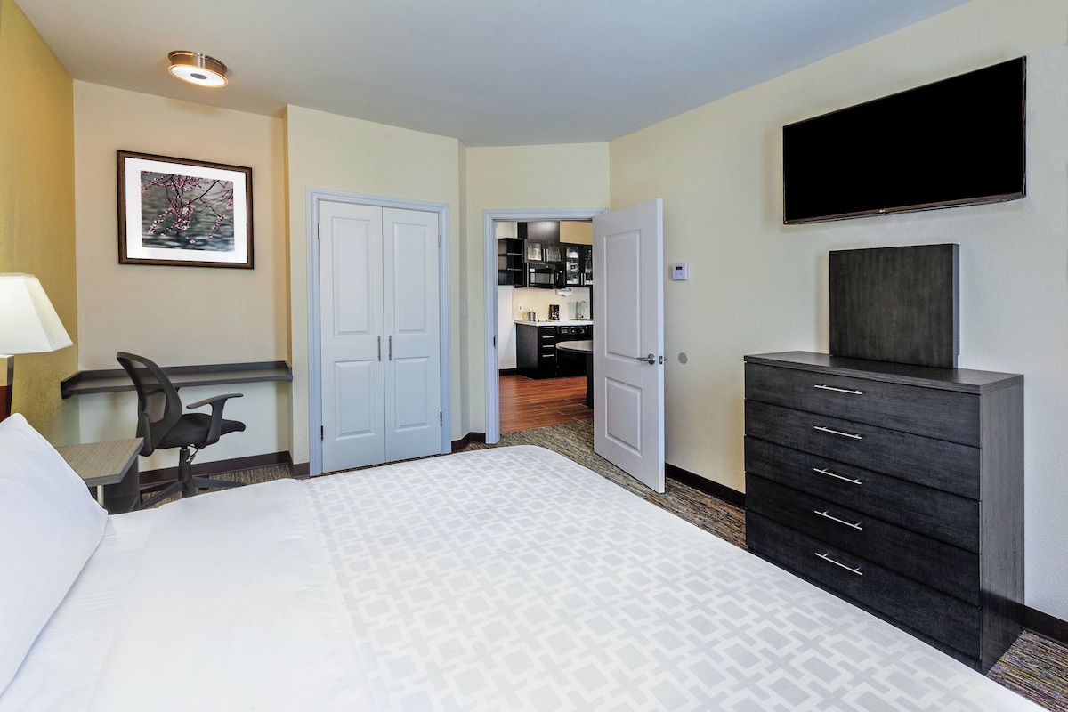 A hotel room with a bed, a desk, a chair, a dresser, a television, and a bathroom visible through an open door.