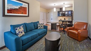 Habitación de hotel con sofá azul, sillón de cuero marrón, mesa de centro redonda, cuadro con paisaje desértico, kitchenette y comedor.