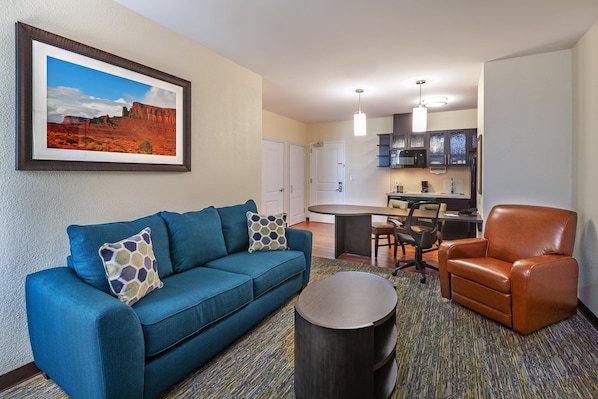 A hotel room with a blue sofa, a brown leather chair, a round coffee table, a framed picture of a desert landscape, a kitchenette, and a dining area.
