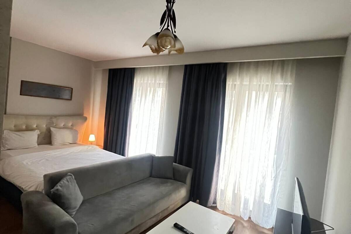 Una habitación de hotel con cama, sofá gris, mesa de centro blanca y un mueble para televisor.