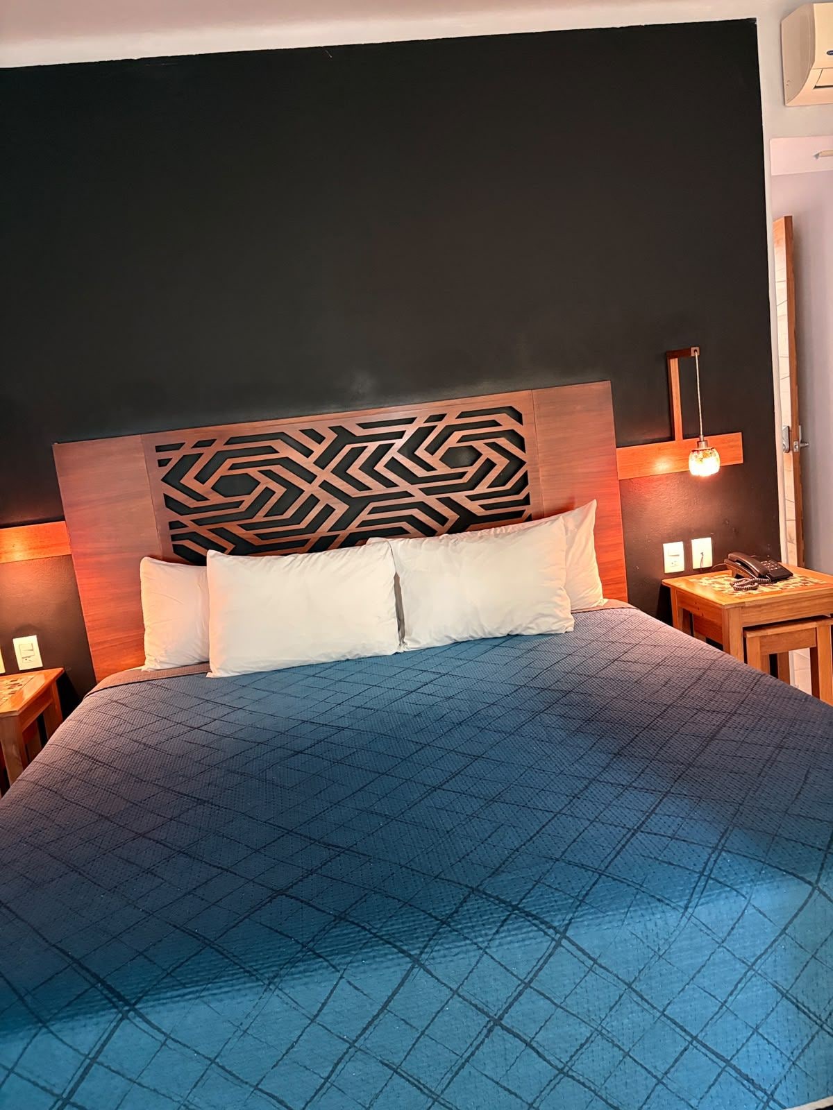 A hotel room with a large bed, a headboard with a geometric pattern, two bedside tables with lamps, and a telephone on one of the tables.