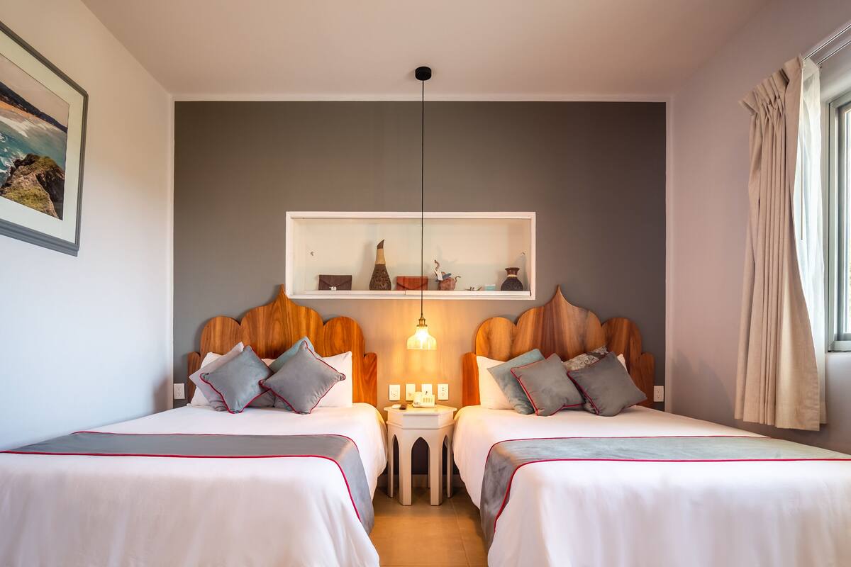 A hotel room with two beds, a small table between them, a shelf above the beds, and a window with curtains.