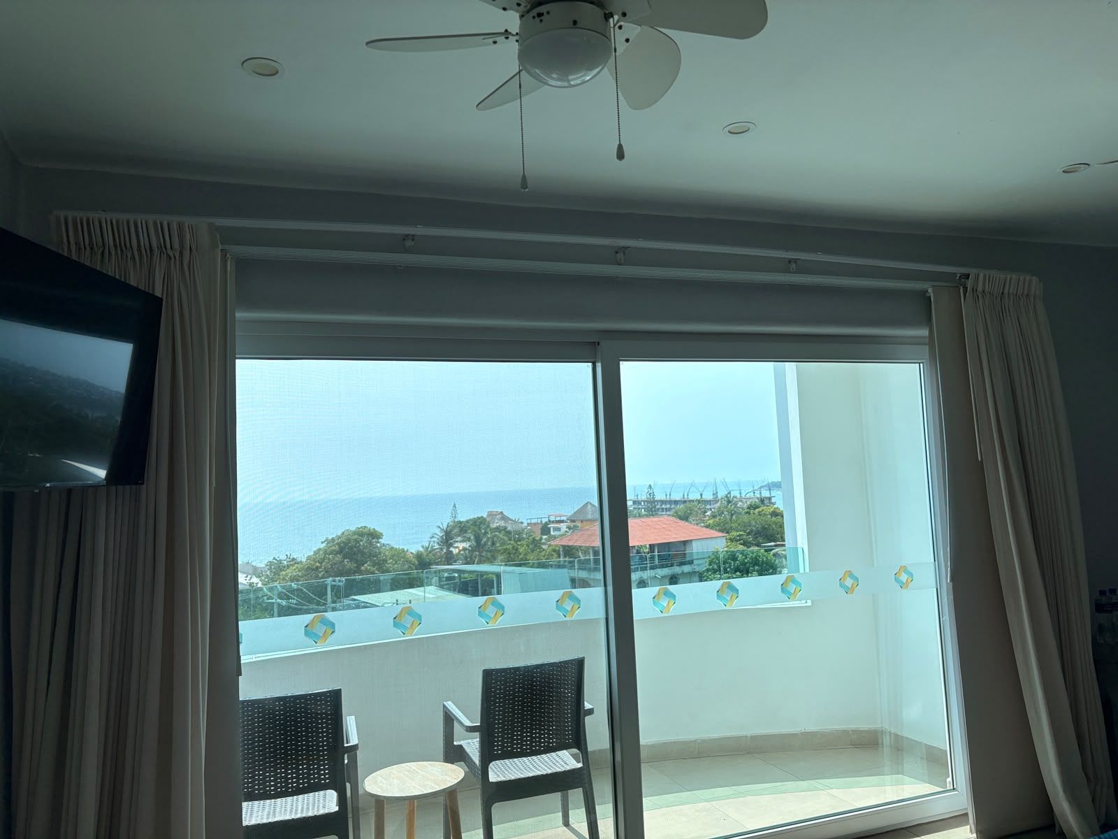 A room with a large sliding glass door leading to a balcony, a ceiling fan, and a flat-screen TV mounted on the wall.