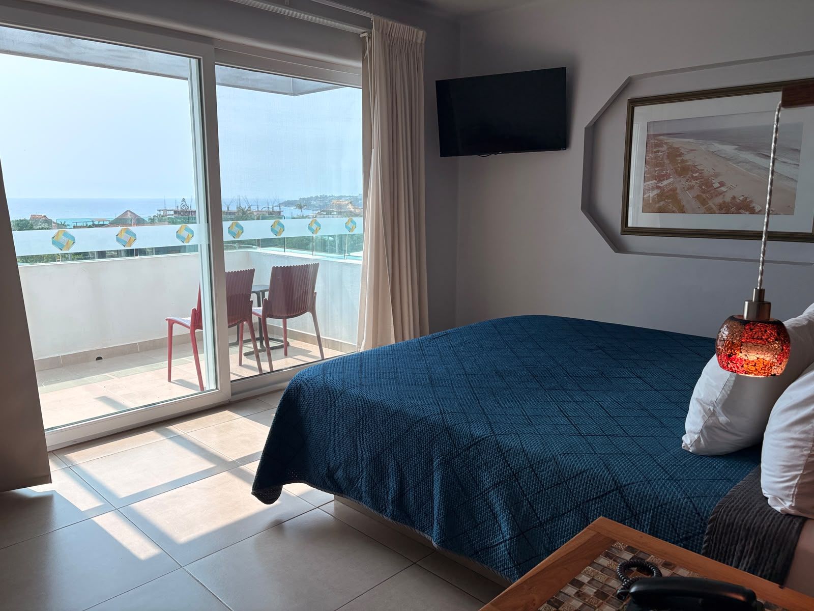 Habitación de hotel con cama, televisor, un cuadro enmarcado, balcón con sillas y vista al mar.