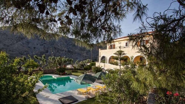 A pool area with a clear view of a building with arches in the background. - Petrinos Milos (Agios Vasileios)