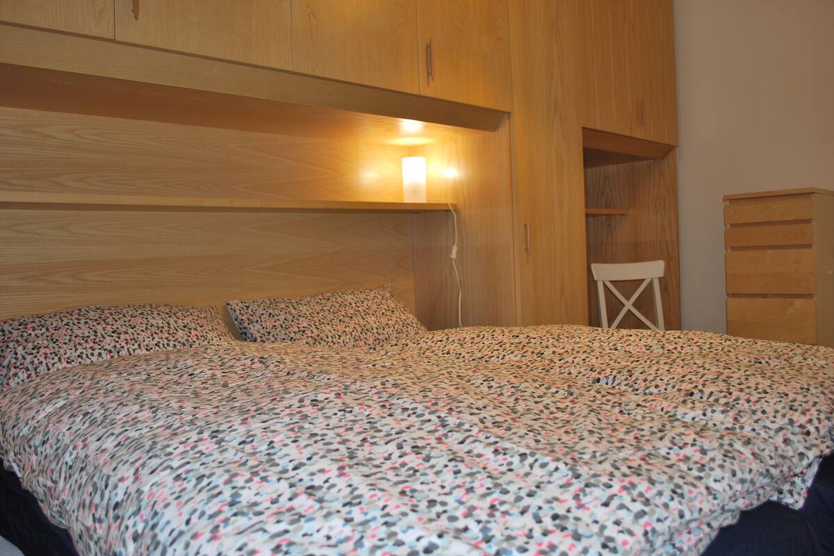 A bedroom with a wooden headboard, a bed with a patterned bedspread, a bedside table, and a chair.