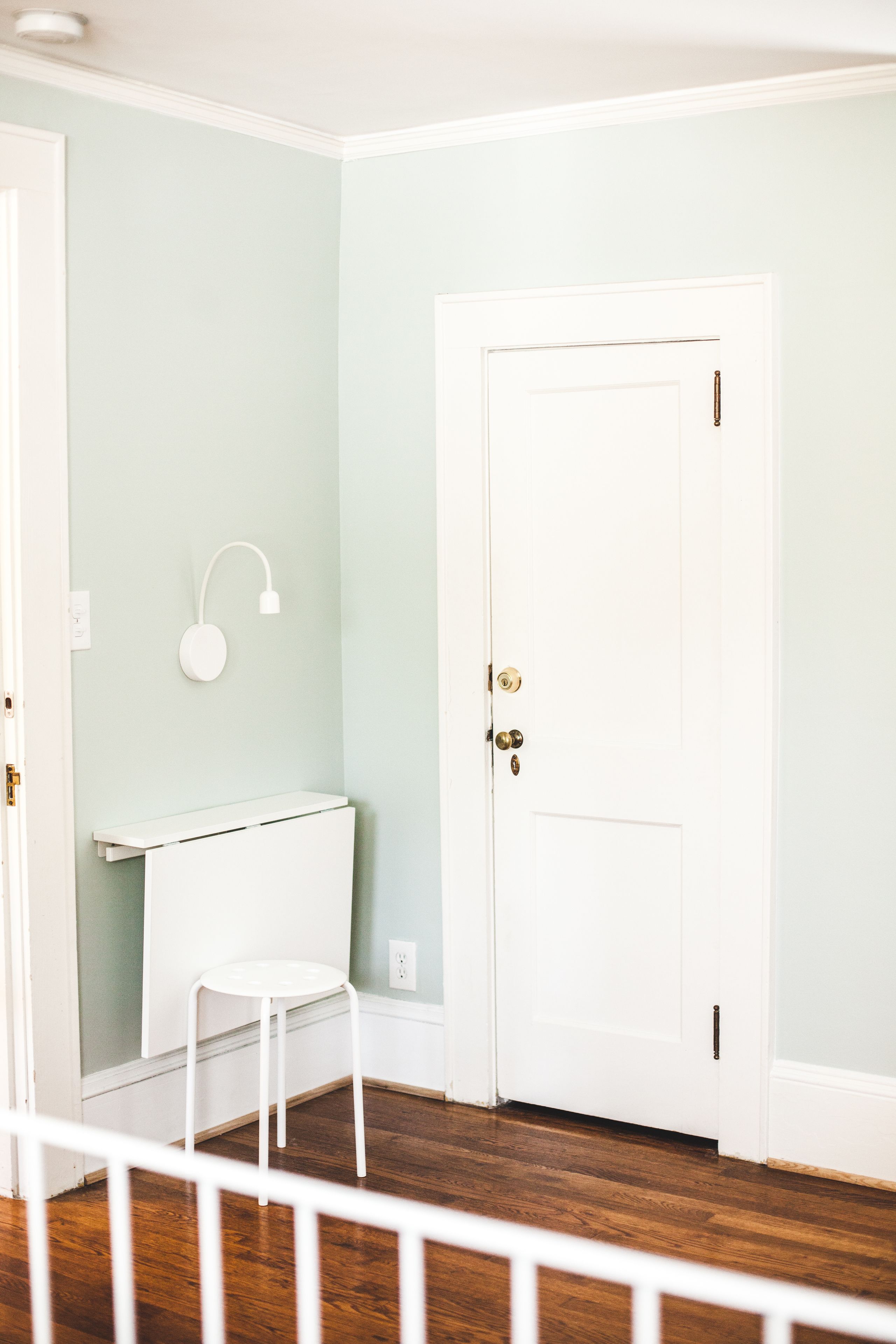 A room with a white door, a small white desk with a chair, and a wall-mounted light fixture.