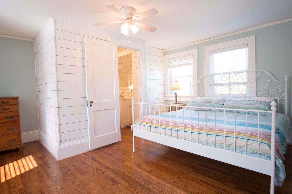 A bedroom with a white metal bed frame, a colorful striped bedspread, a ceiling fan, and a wooden dresser.