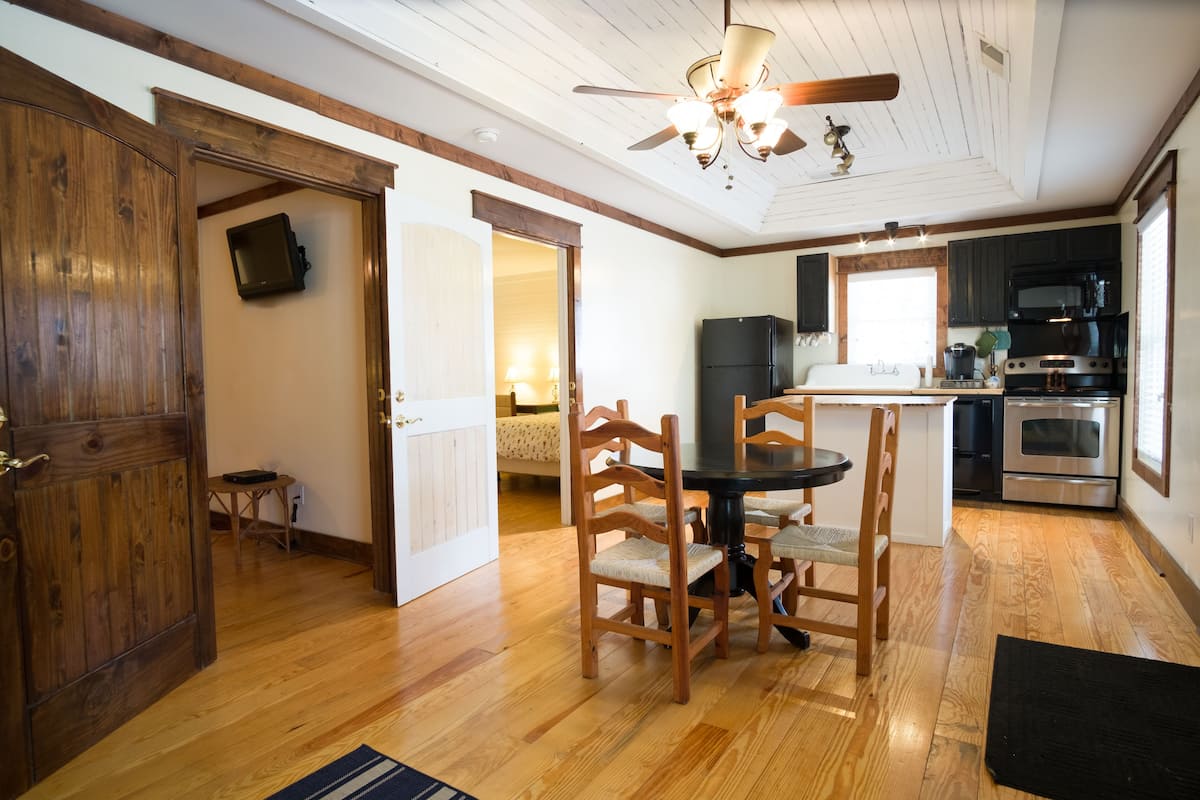 A spacious kitchen with wooden flooring, a dining area with a round table and chairs, and a ceiling fan.