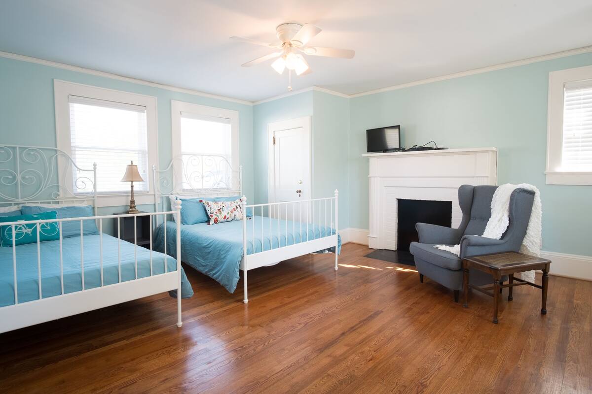 A bedroom with a white metal bed frame, a blue bedspread, a grey armchair, a small wooden table, a white fireplace, and a ceiling fan.