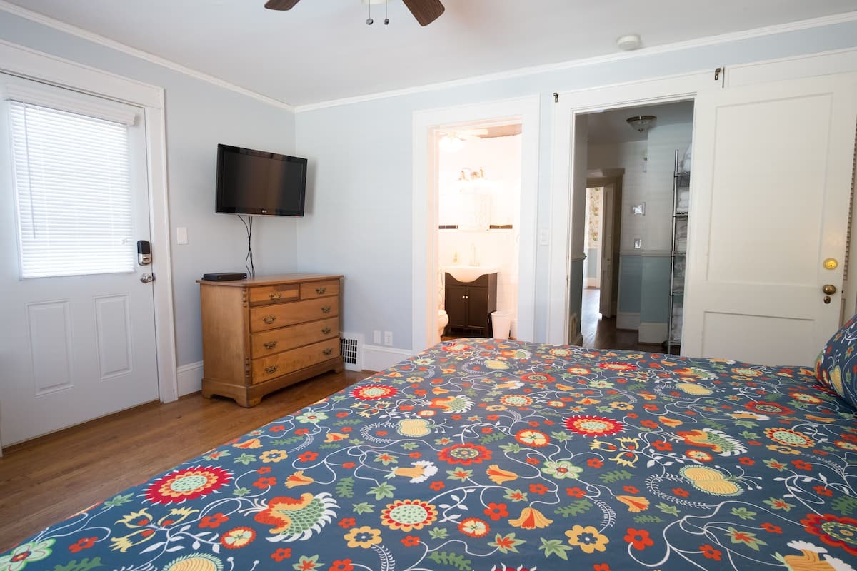 A bedroom with a floral bedspread, a wooden dresser, a television on the wall, and a door leading to a bathroom.