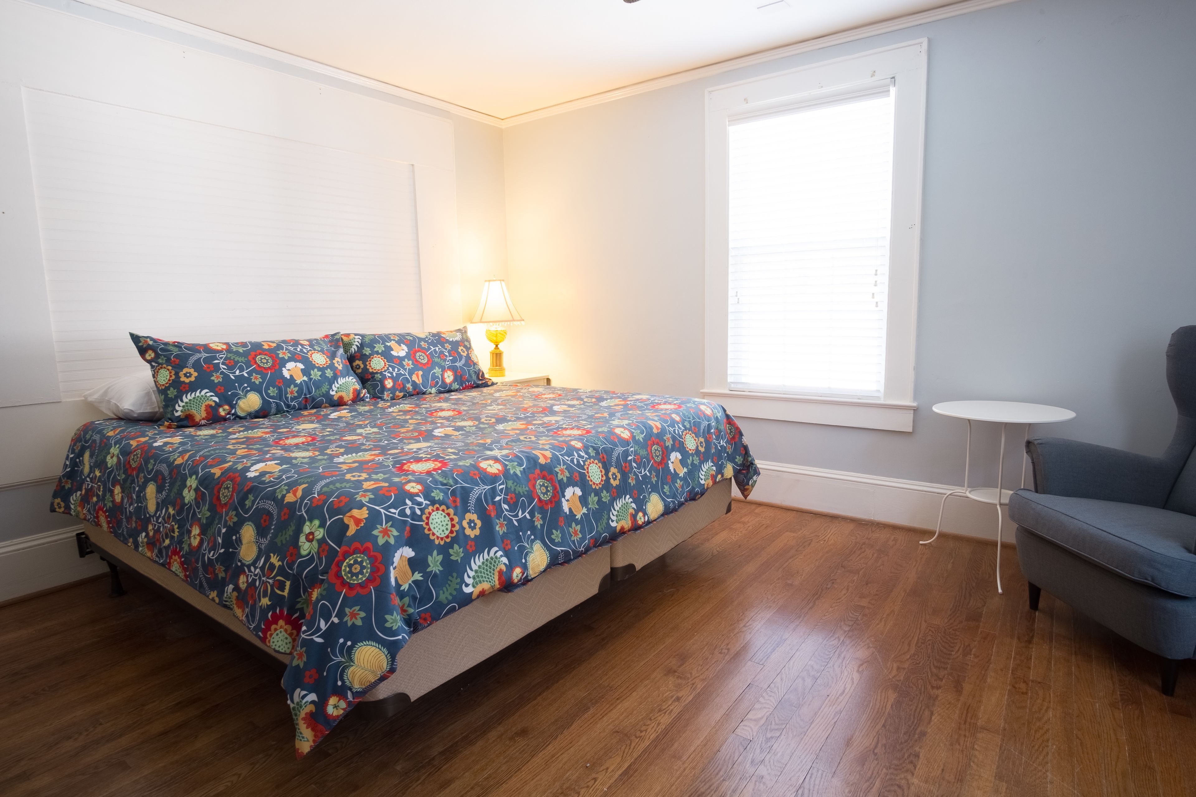 Un dormitorio con cama, mesita de noche, lámpara, silla y ventana.