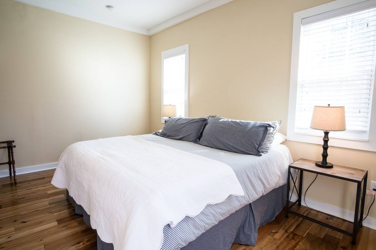 A bedroom with a large bed, a bedside table with a lamp, and a window with blinds.