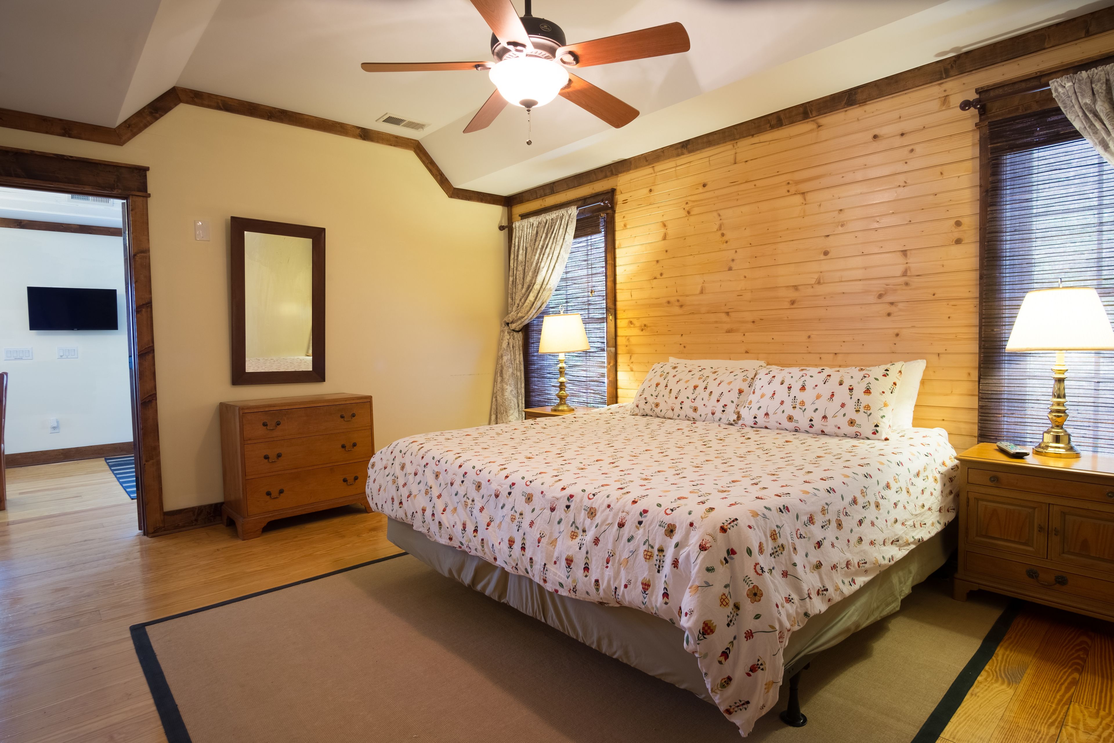 Un dormitorio con ventilador de techo de madera, cama con ropa de cama floral, mesita de noche con lámpara y cómoda.